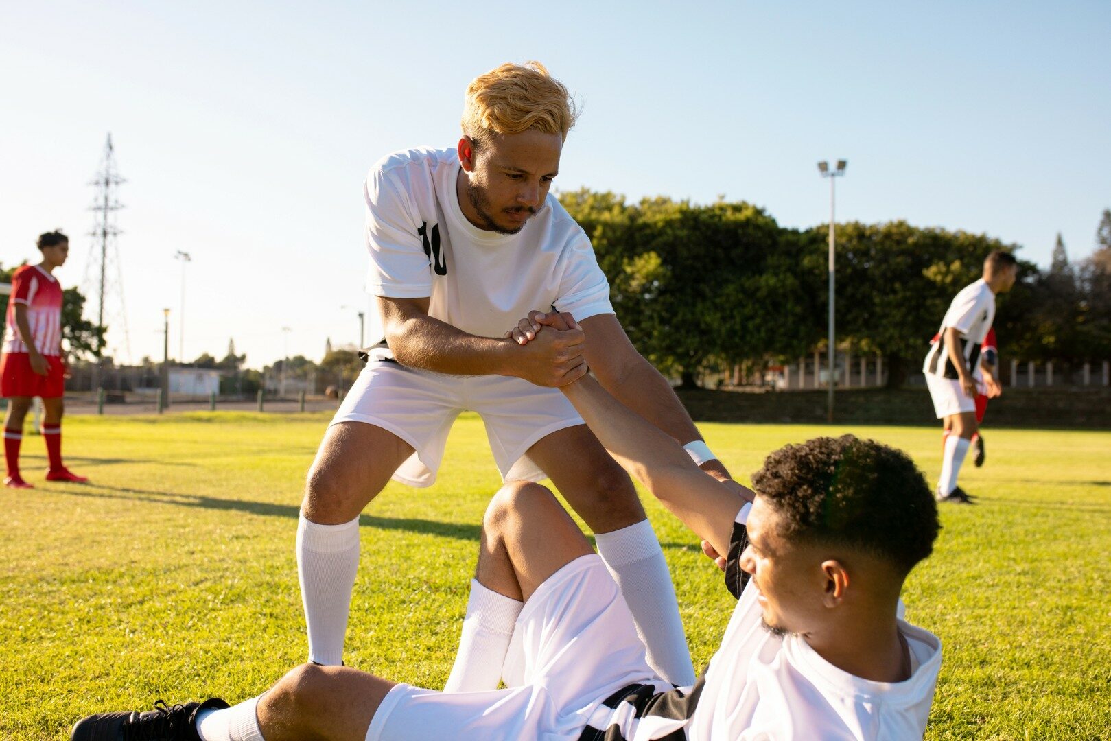 Blanke speler geeft hand aan gewonde teamgenoot liggend op het veld in het opstaan tegen de heldere hemel.